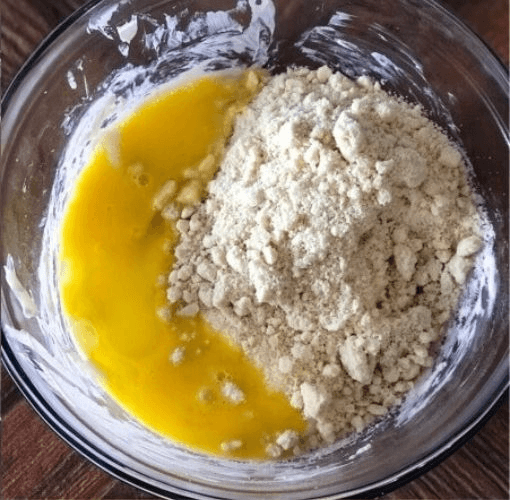 Beaten egg and almond flour resting on top of melted cheese in a glass mixing bowl, before being stirred together