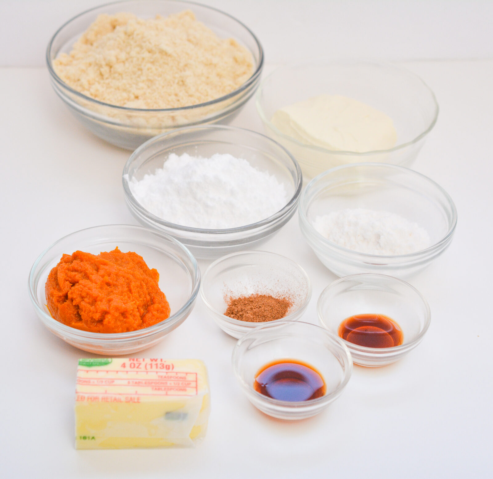 Glass bowls of almond flour, cream cheese and two white powdered ingredients, with smaller bowls of pumpkin puree, ground spice and vanilla, and a wrapped stick of butter in front