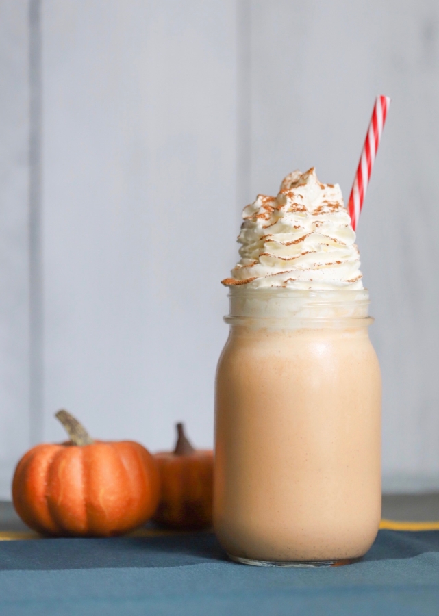keto pumpkin cheesecake shake with whip cream on top and a straw sticking out. Two small pumpkin figurines beside the shake