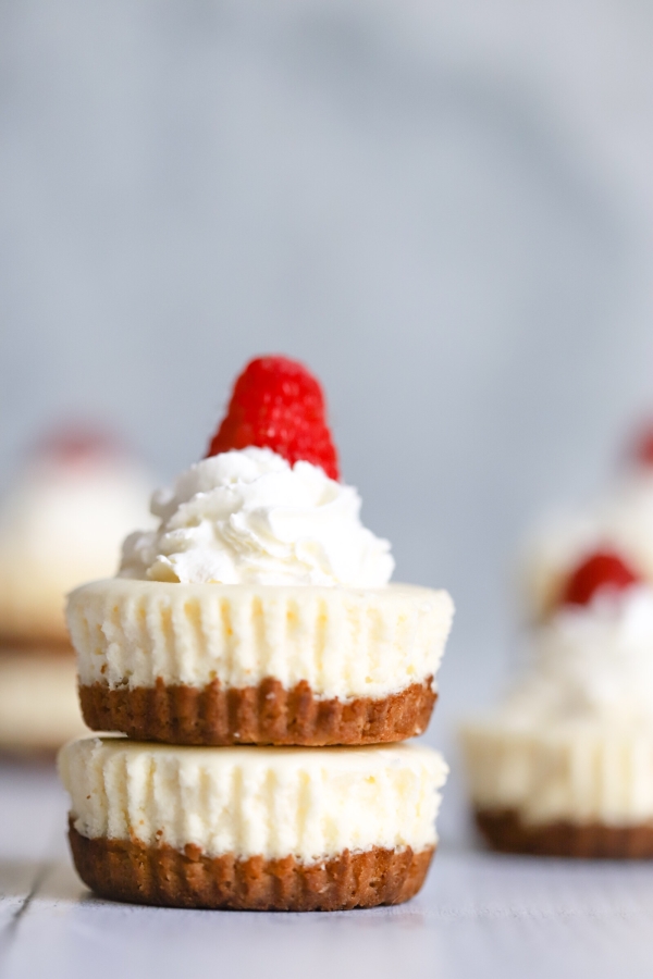 two mini cheesecakes on top of each other with a little whip cream and raspberry on top. More mini cheesecakes in the background