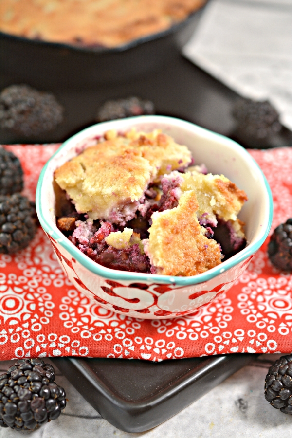 front view of keto cobbler in a bowl with black berries scattered next to it and left over of the cobbler in a skillet in the background