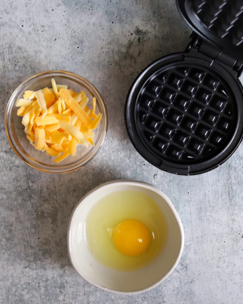 top view of a bowl of cheese, a bowl with an egg, and a mini waffle maker on the side