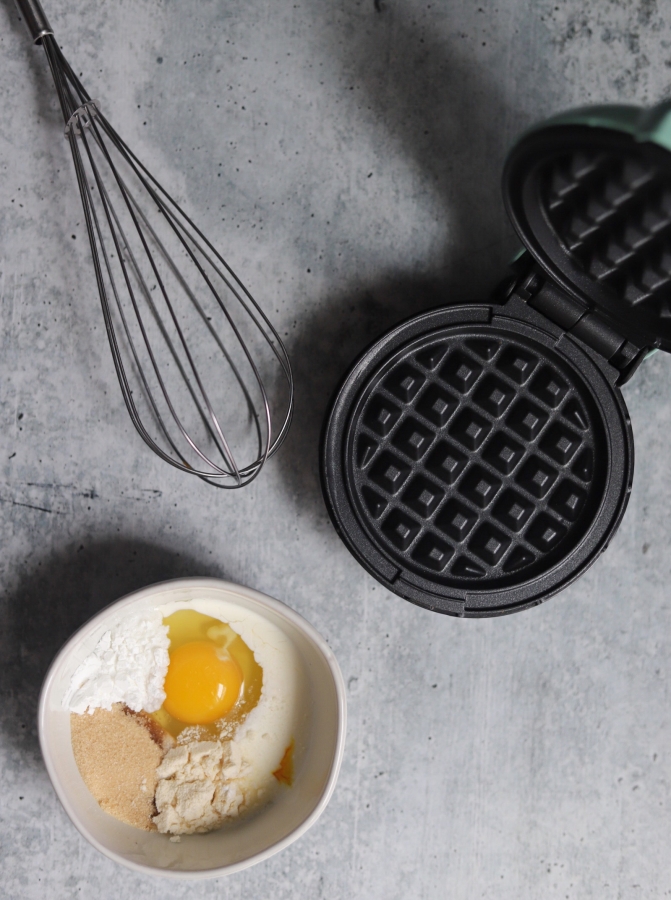 top view of the ingredients in a bowl, a whisk, and the mini waffle griddle