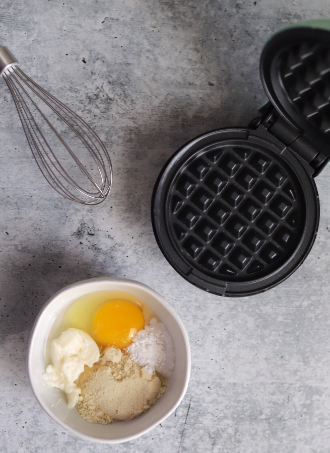top view shot of ingredients in a bowl, mini waffle maker, and a whisk