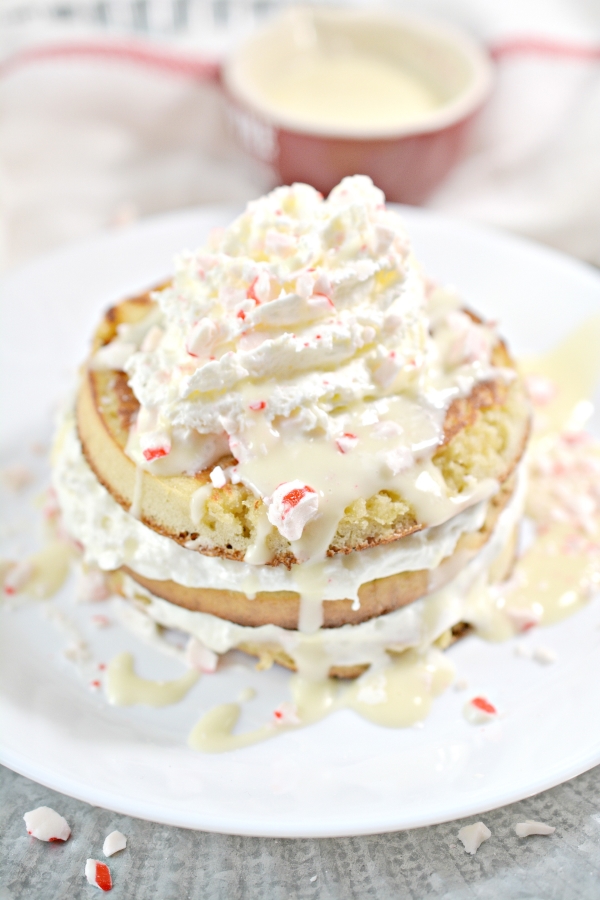 stack of keto peppermint pancakes on a plate with drizzled white chocolate, whipped cream, and crushed peppermint candies on top