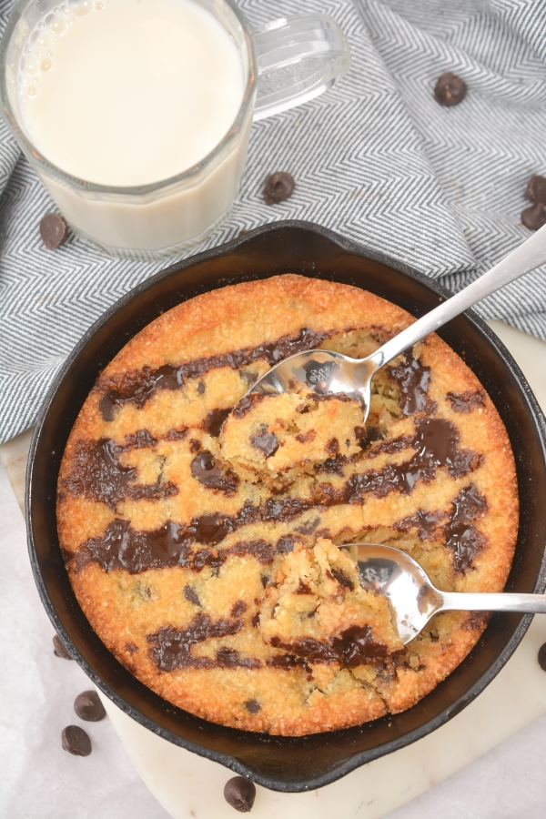 top view of cookie in a skillet with two spoons dug into the keto chocolate chip skillet cookie and a glass of milk on the side.